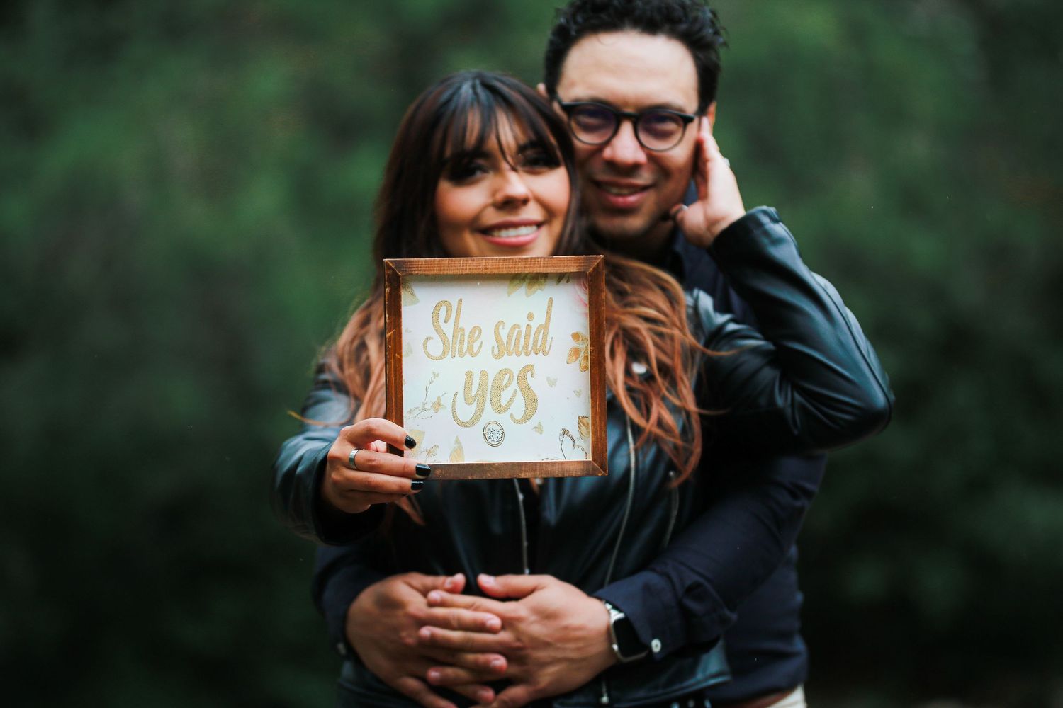 A happy couple embraces while holding a She Said Yes engagement announcement sign.