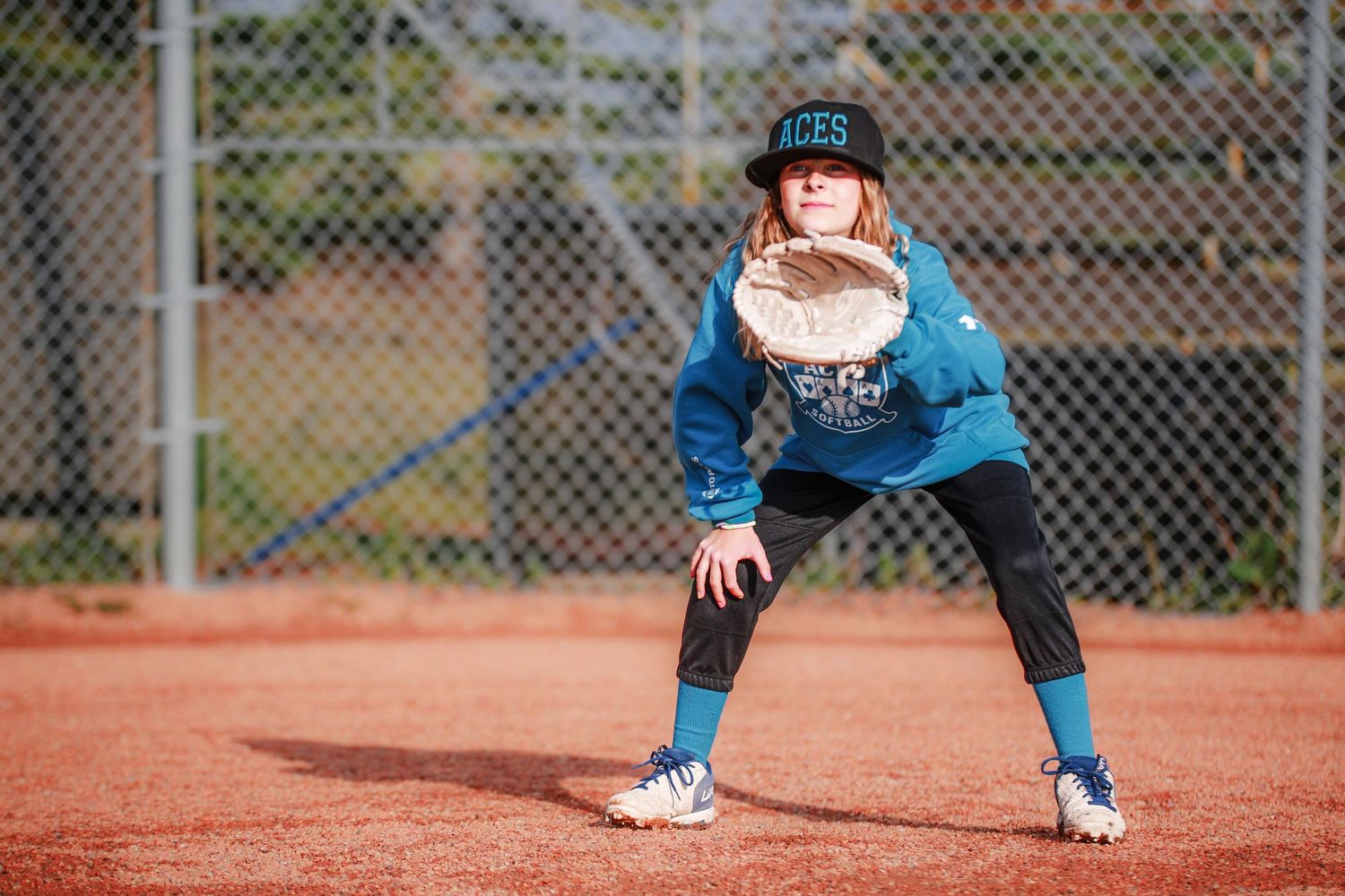 A young softball player in a blue Aces uniform crouches in fielding position on a dirt diamond.