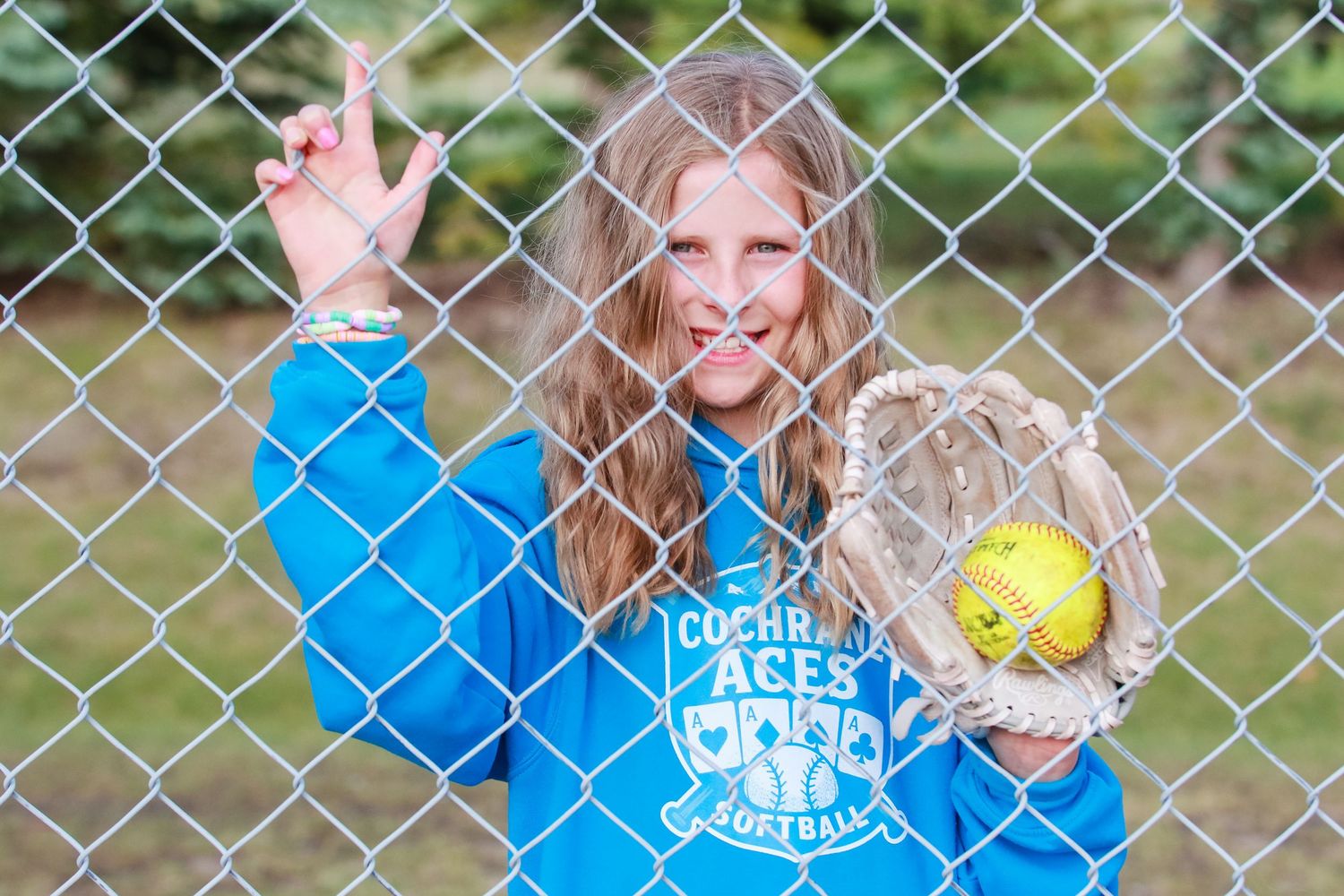 A smiling girl in a blue Cochrane Aces softball hoodie holds a glove and ball behind a chain-link fence.