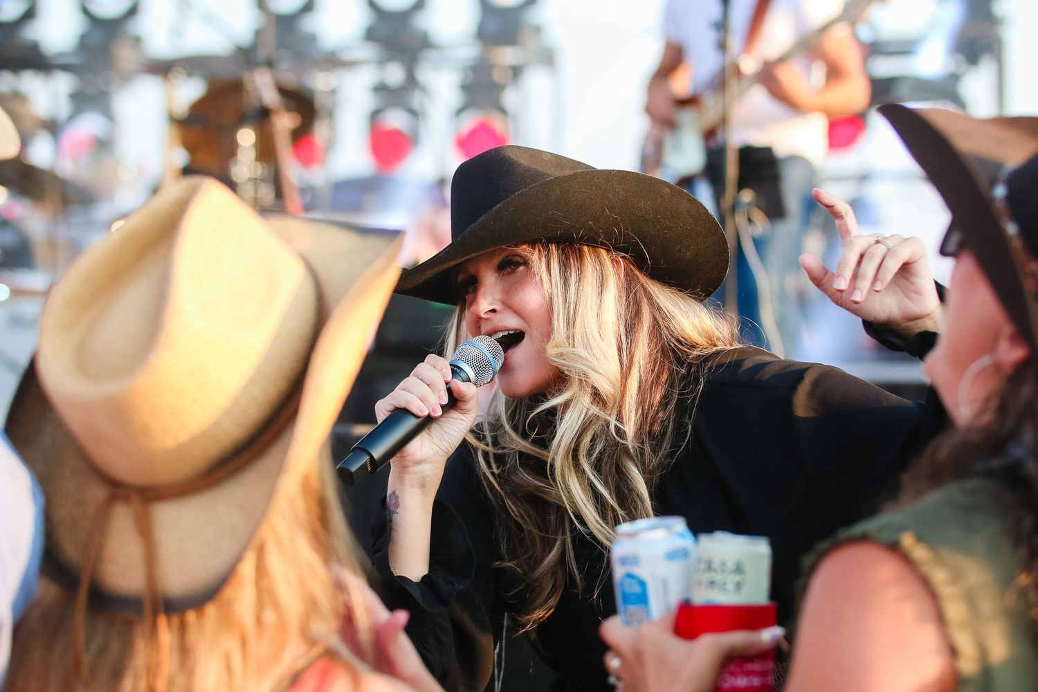 A woman in a black cowboy hat sings into a microphone at an outdoor country music festival.