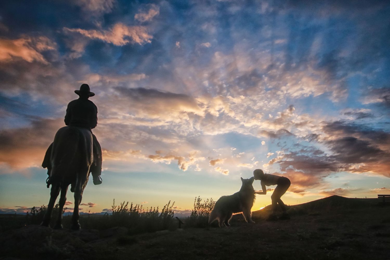 Cowboy on horseback and person with dog silhouetted against dramatic colorful sunset sky.