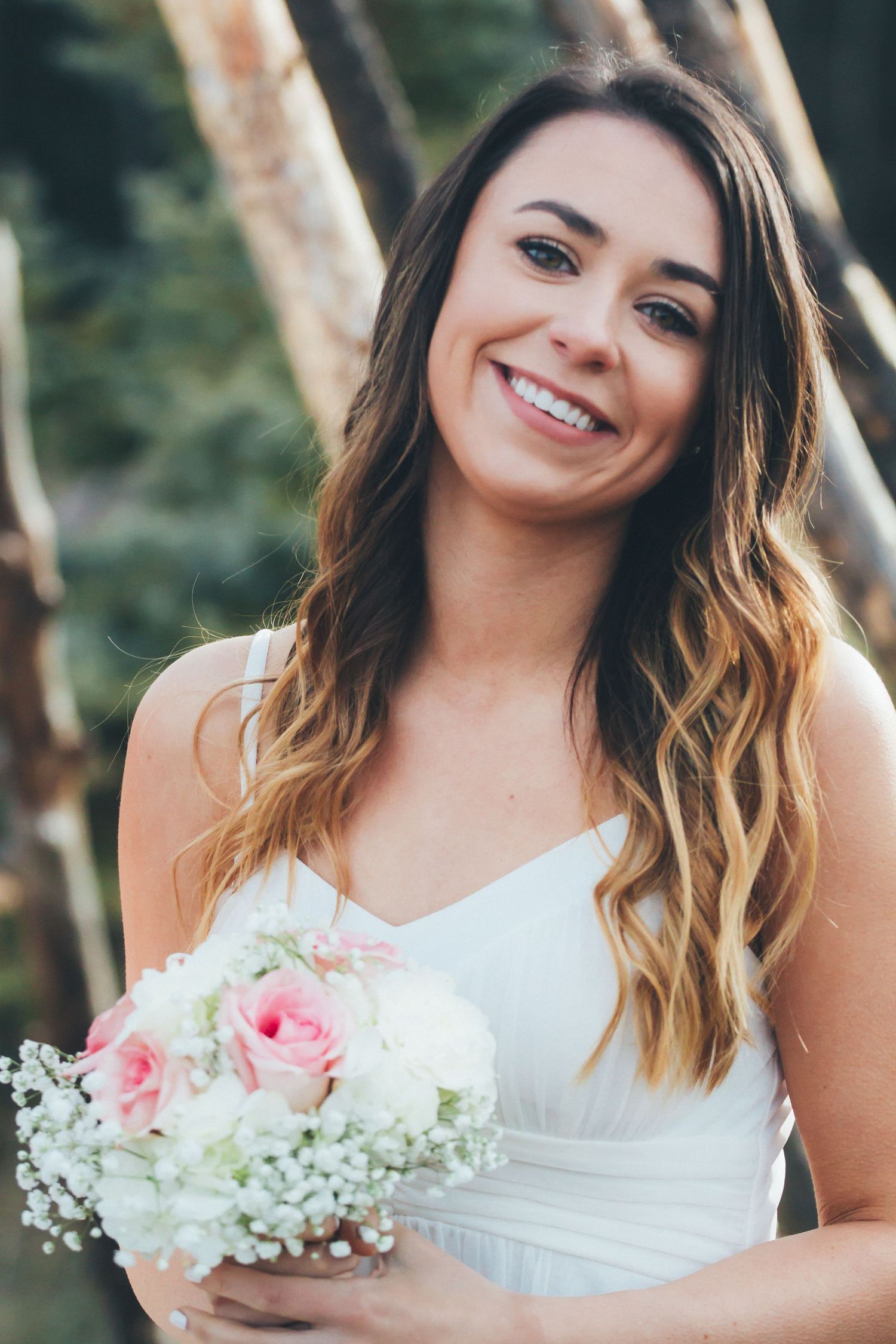 A smiling bride in a white dress holds a bouquet of pink and white roses outdoors.