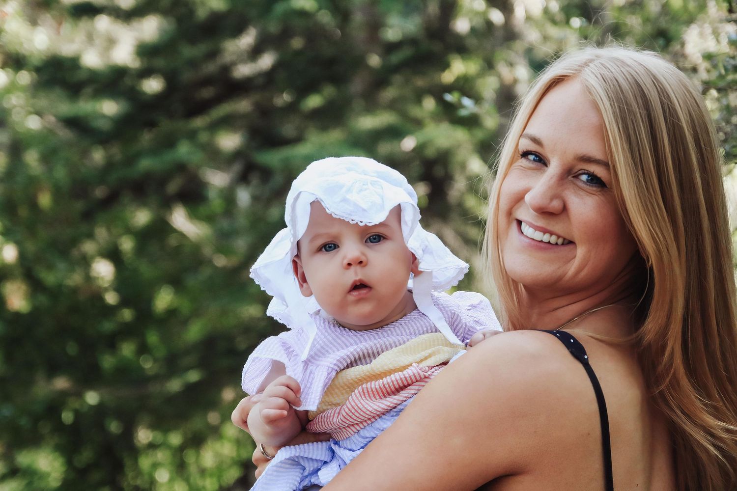 Smiling blonde woman holding an adorable baby wearing a white sun hat outdoors.