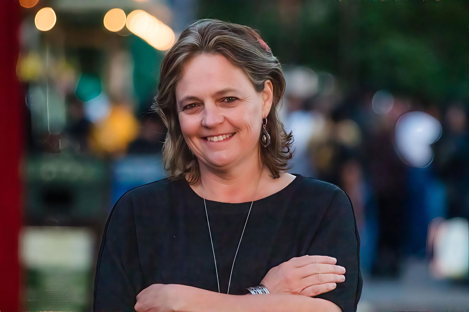 Smiling woman with short hair standing outdoors at a lively evening event with arms crossed.