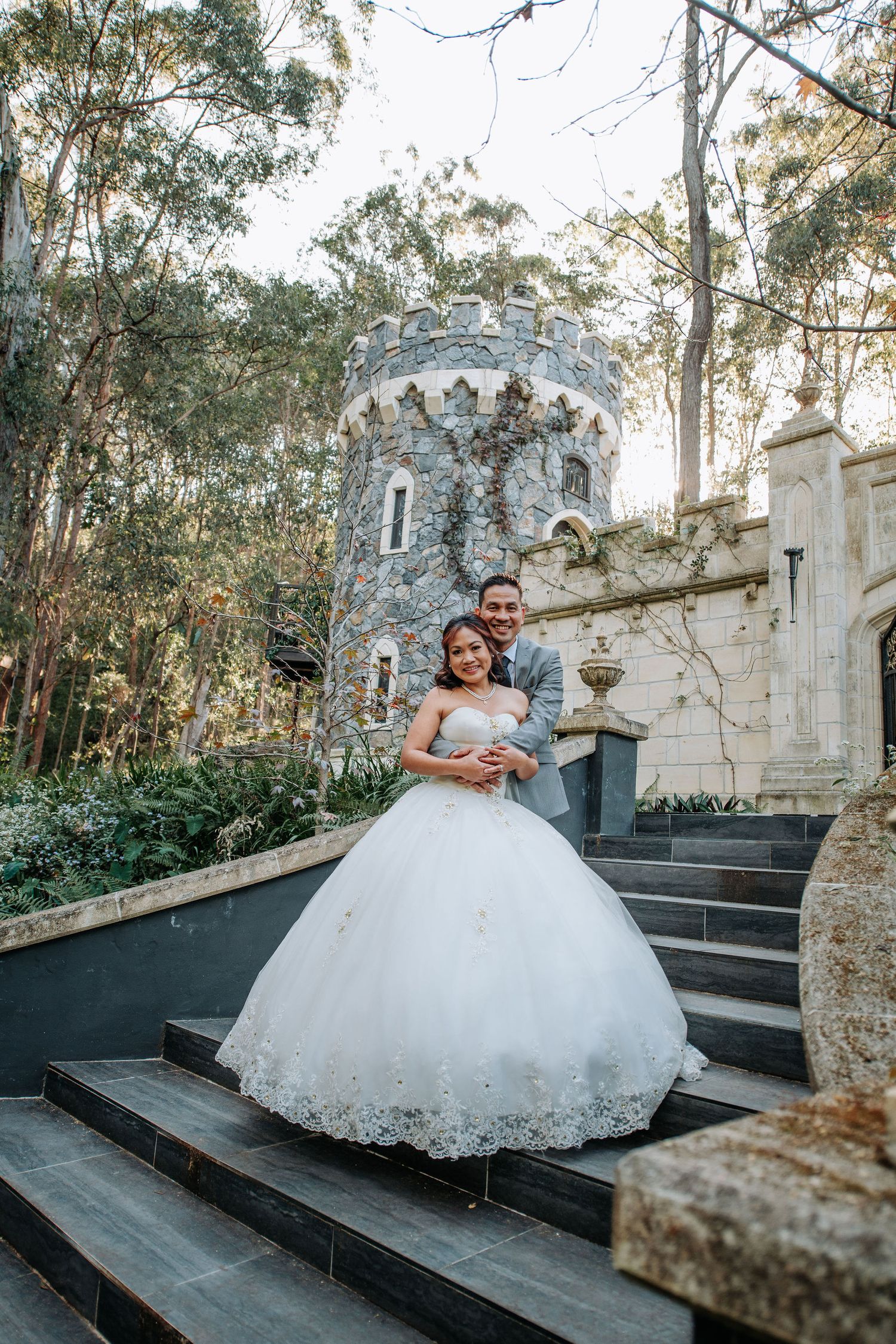 A wedding portrait on stone steps with a fairytale castle tower backdrop in a forest setting.