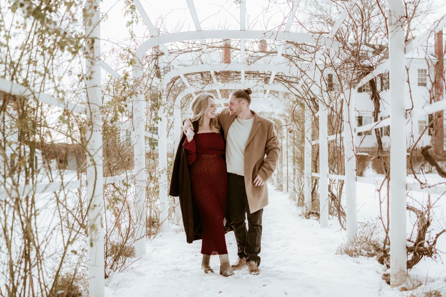 Couple walking under snow-covered white pergola arch in winter, wearing elegant coats in red and tan, surrounded by bare vines.