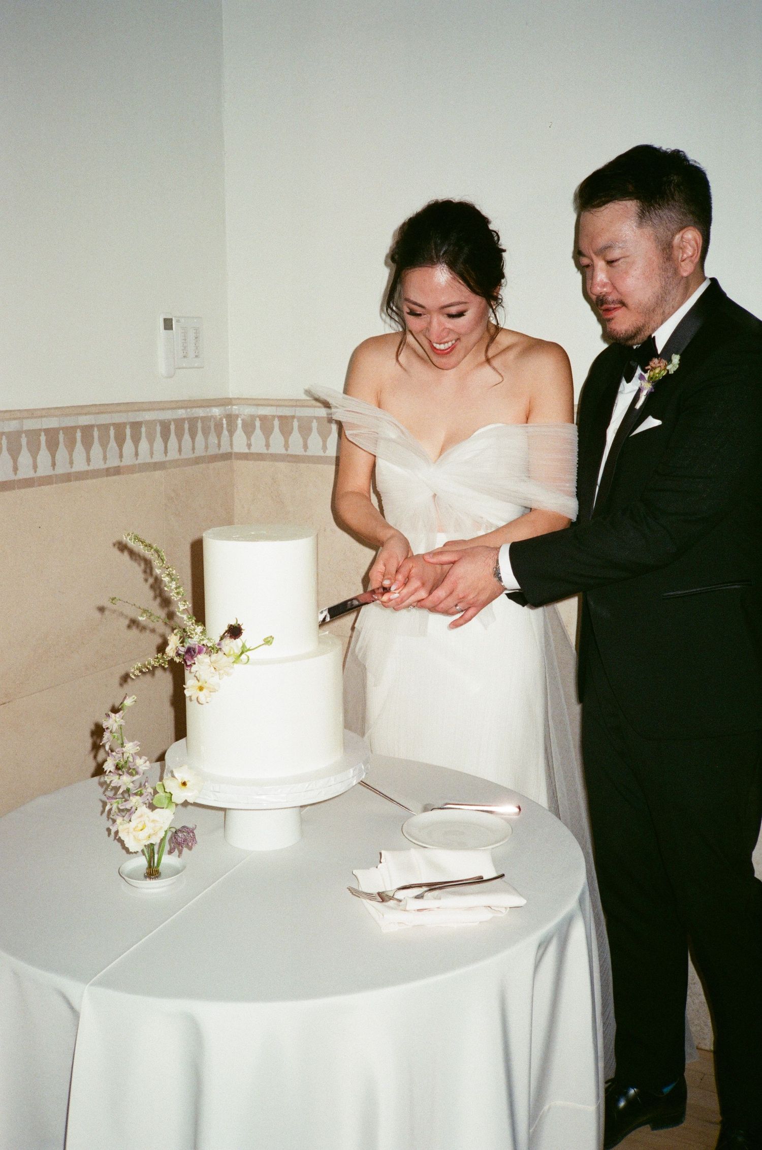 A couple in formalwear cuts into their elegant two-tier wedding cake at a wedding reception.