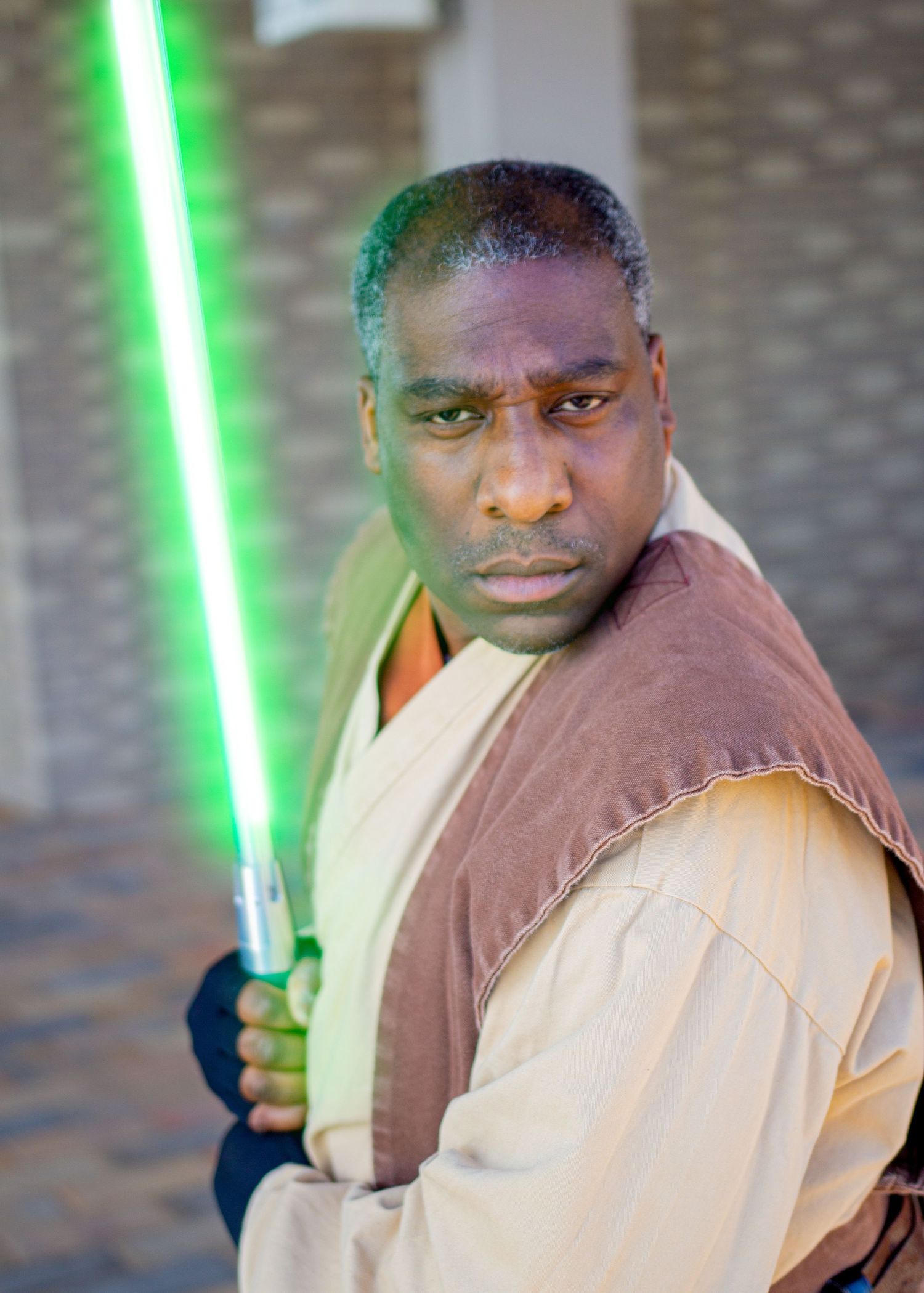 Star Wars cosplayer in Jedi robes poses with green lightsaber against brick wall.