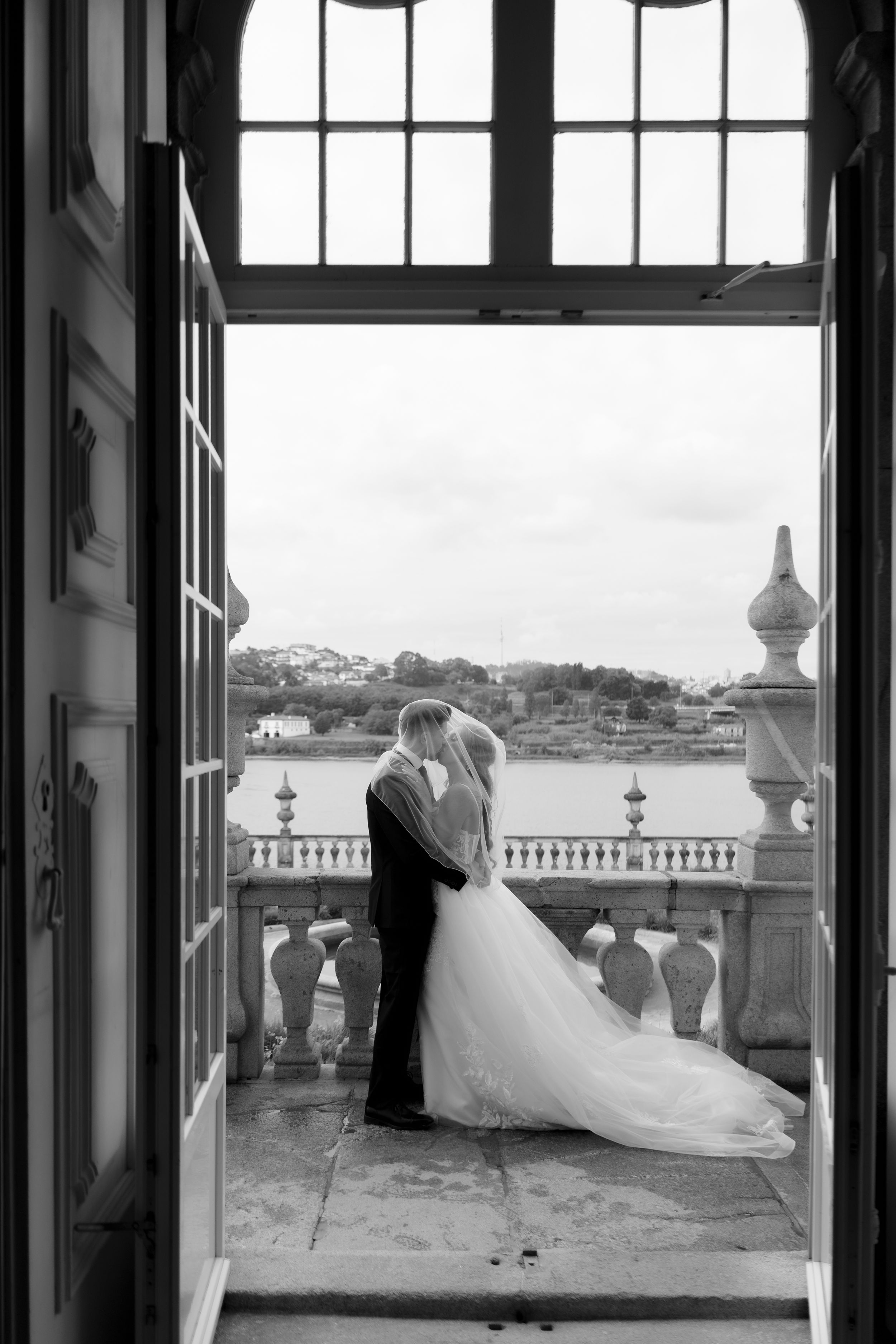 Black and white photo of bride and groom kissing on ornate balcony overlooking river.