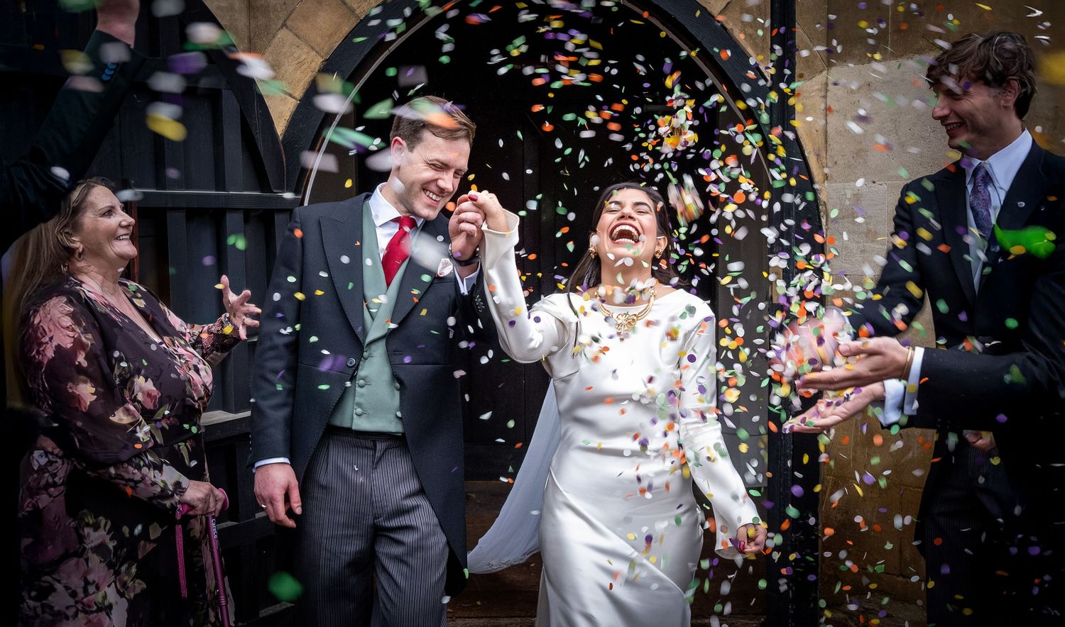 Newlyweds walk through confetti shower outside brick church surrounded by celebrating guests.