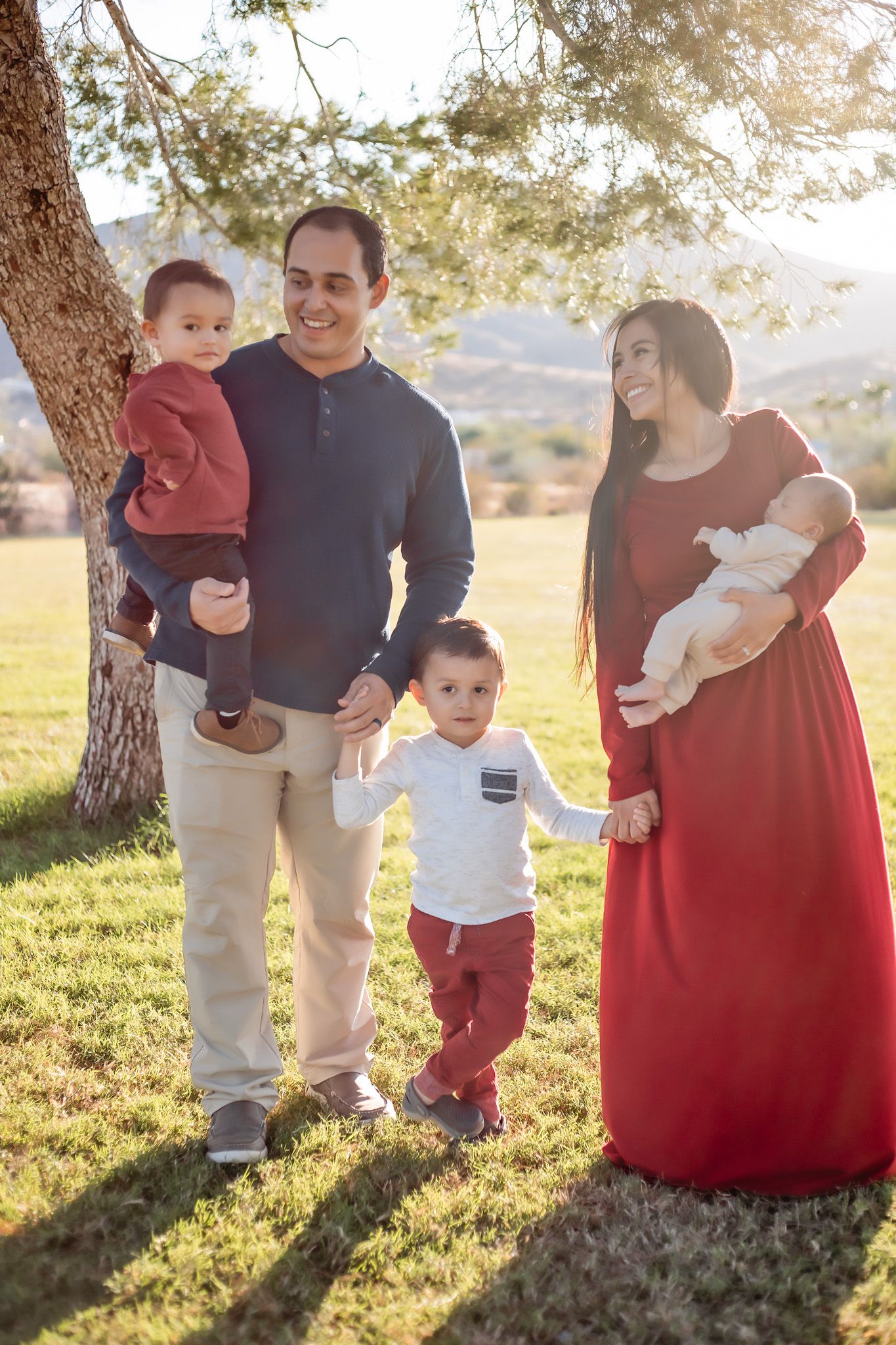 Dad, mom, and 3 boys smiling together outside in the sun under a shaded tree