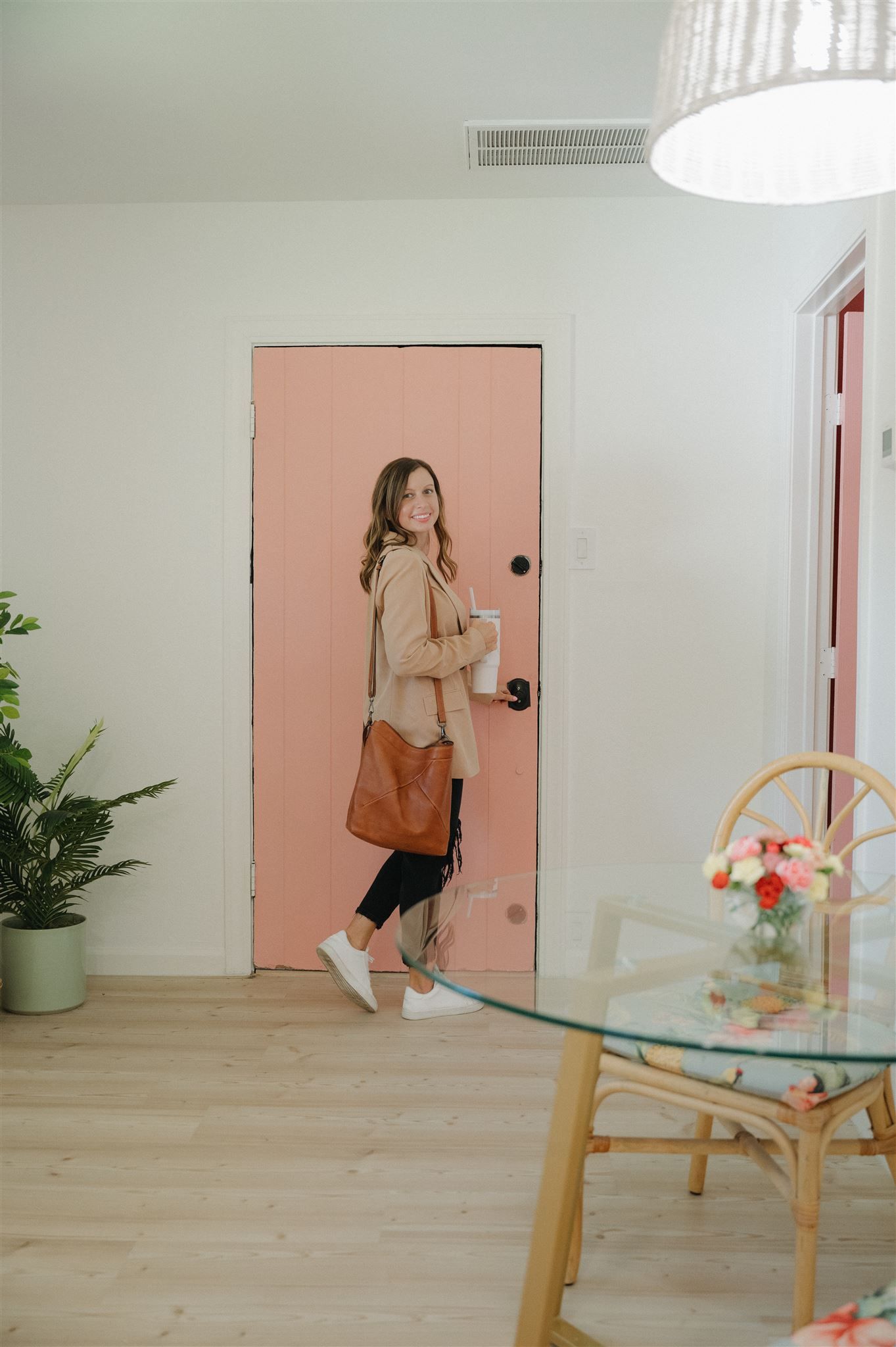 Someone in a beige blazer poses in front of a pink door holding a brown leather bag.