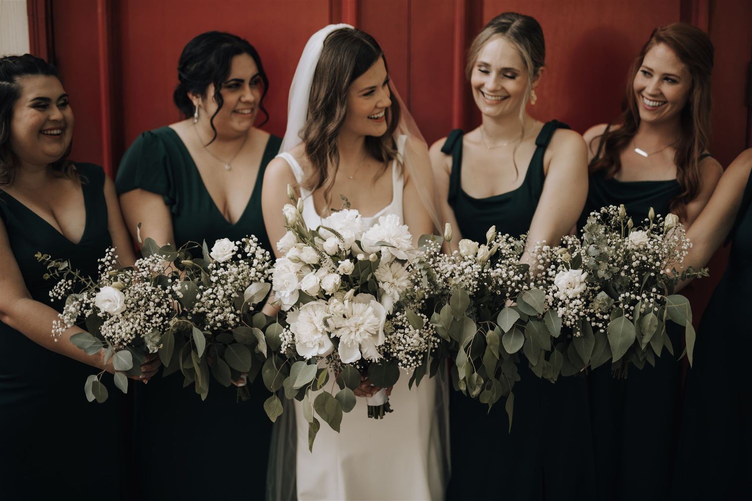 Bridesmaids in dark green dresses hold lush white and green bouquets while sharing a candid moment.