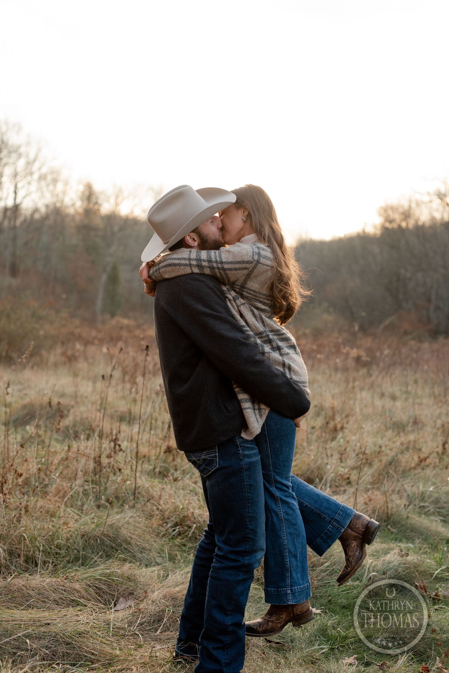 A couple shares a romantic kiss while standing in a sunlit field at sunset.