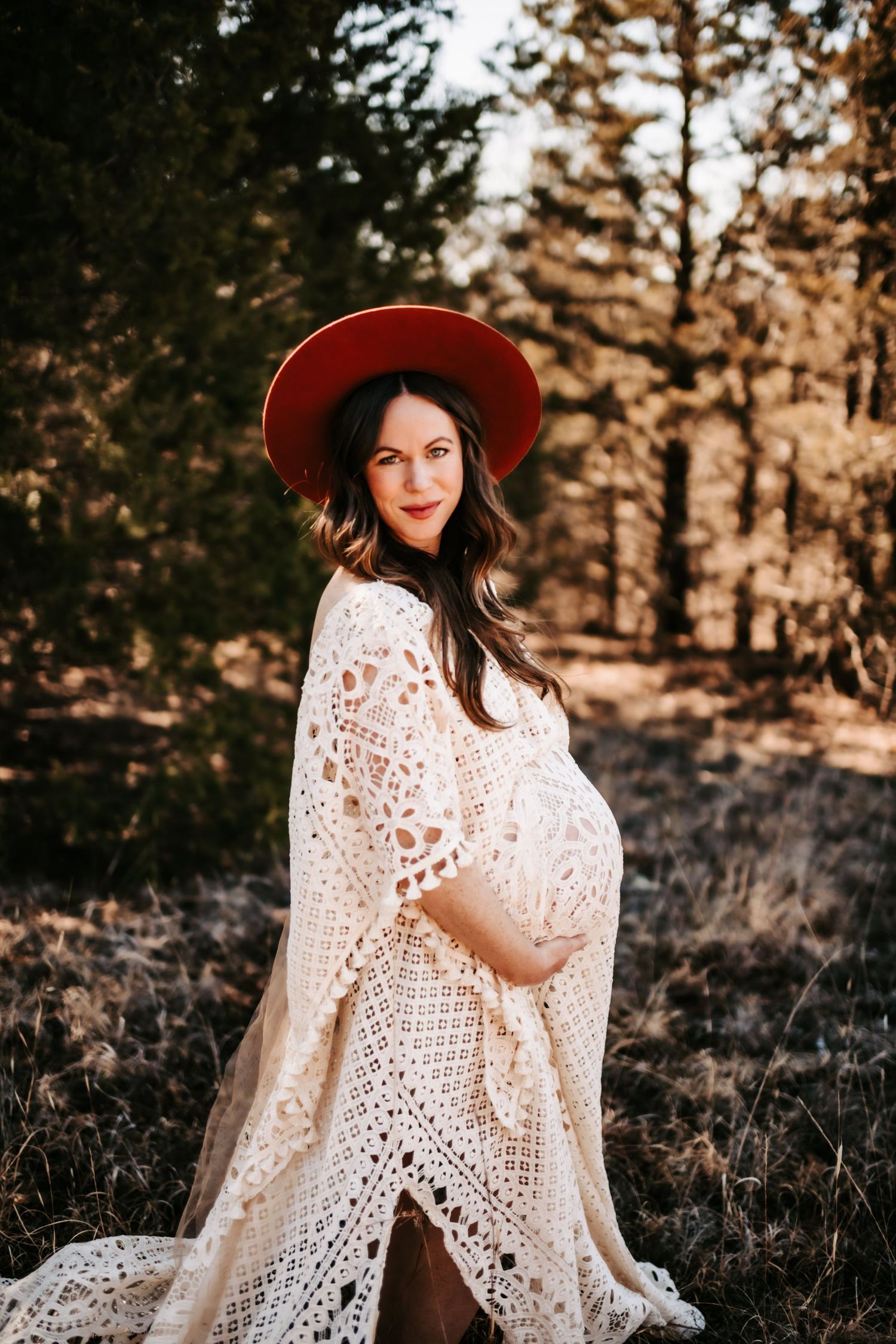 A dreamy maternity photo features a flowing lace dress and red hat in a natural outdoor setting.