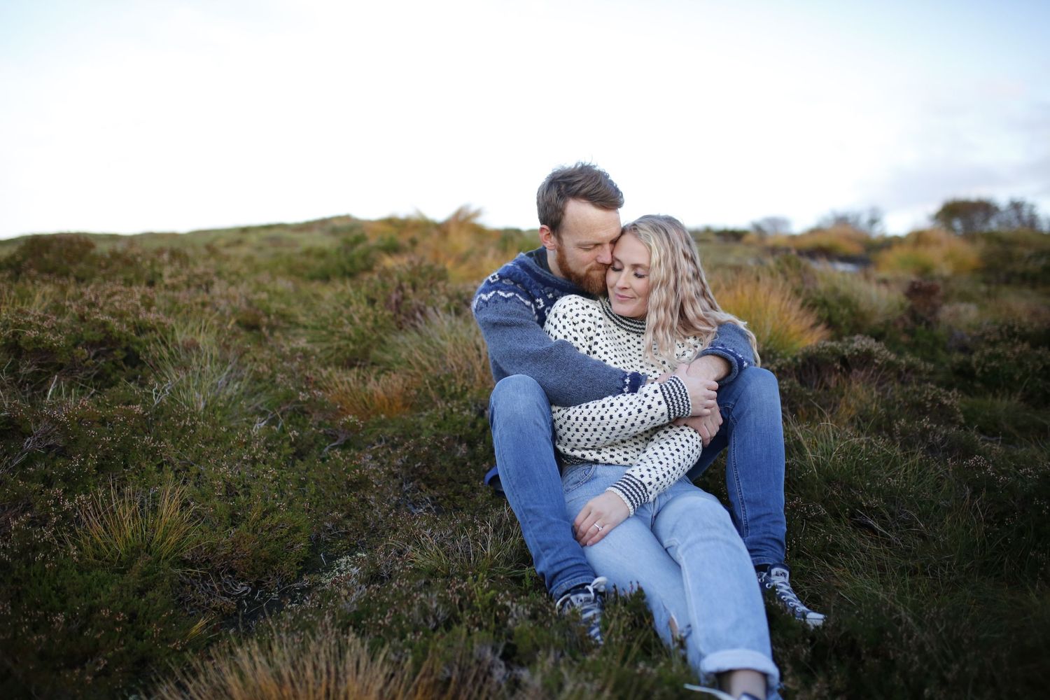 A couple shares an intimate moment while sitting together on a grassy hillside at sunset.