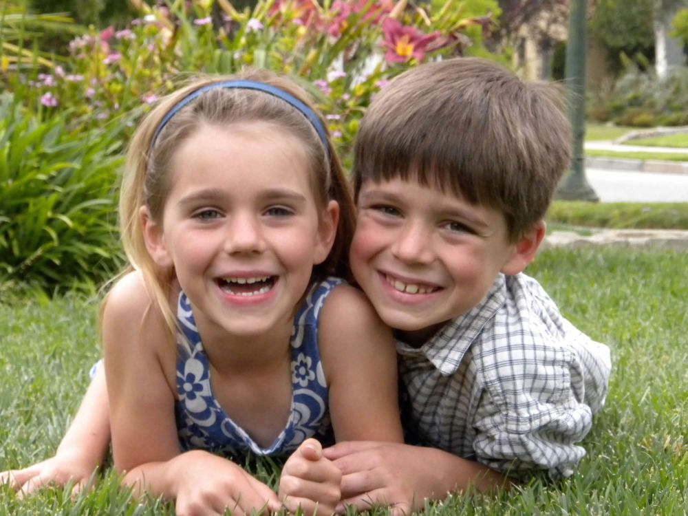 Two children lying in grass with colorful flowers blooming in background.