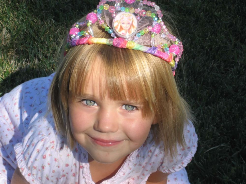 Young child wearing a colorful princess tiara and white dress smiling at camera outdoors.