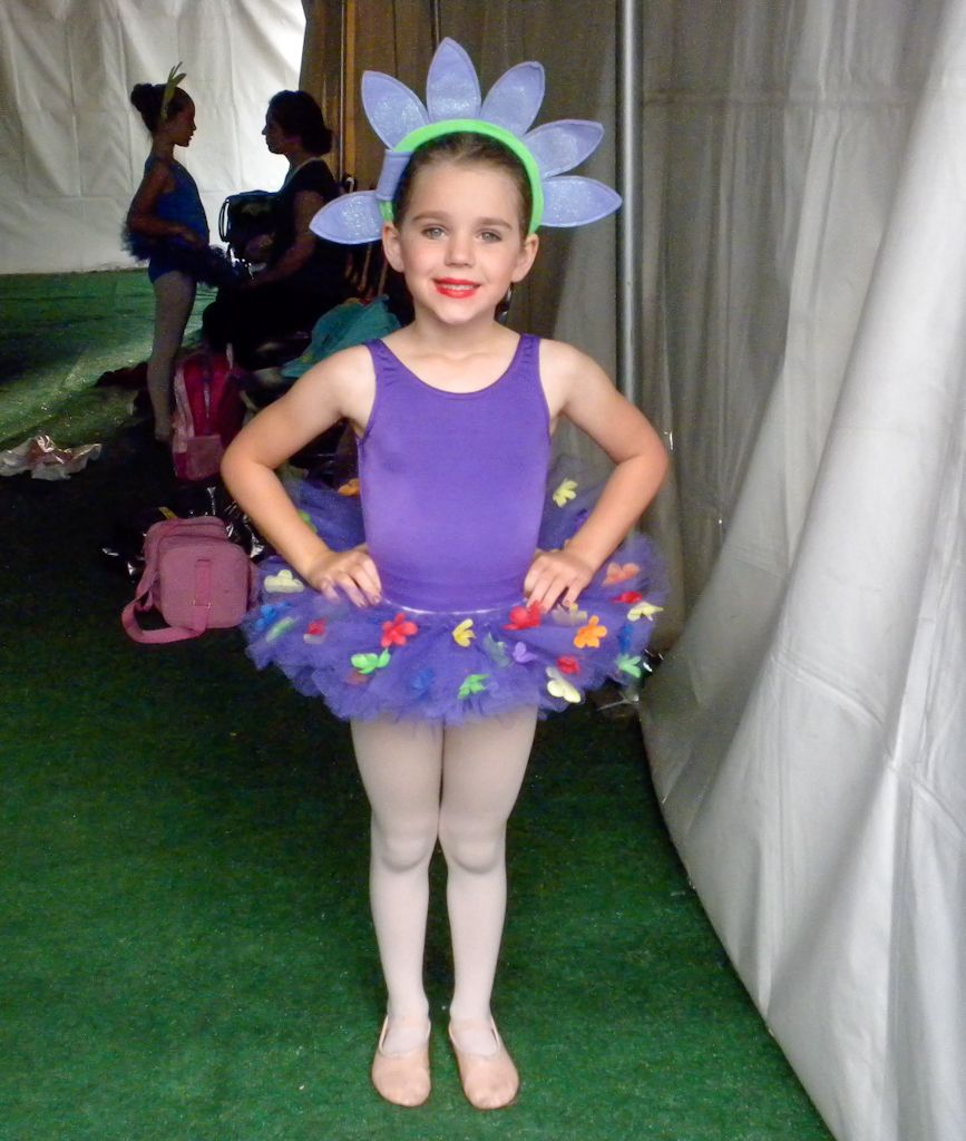 Young dancer in a purple tutu with flower headpiece poses on green carpet at dance recital.