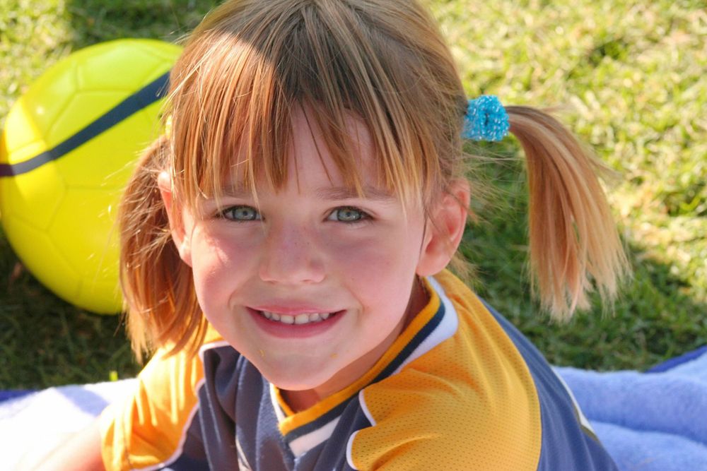 Child in yellow sports uniform smiling while lying on blue blanket outdoors.