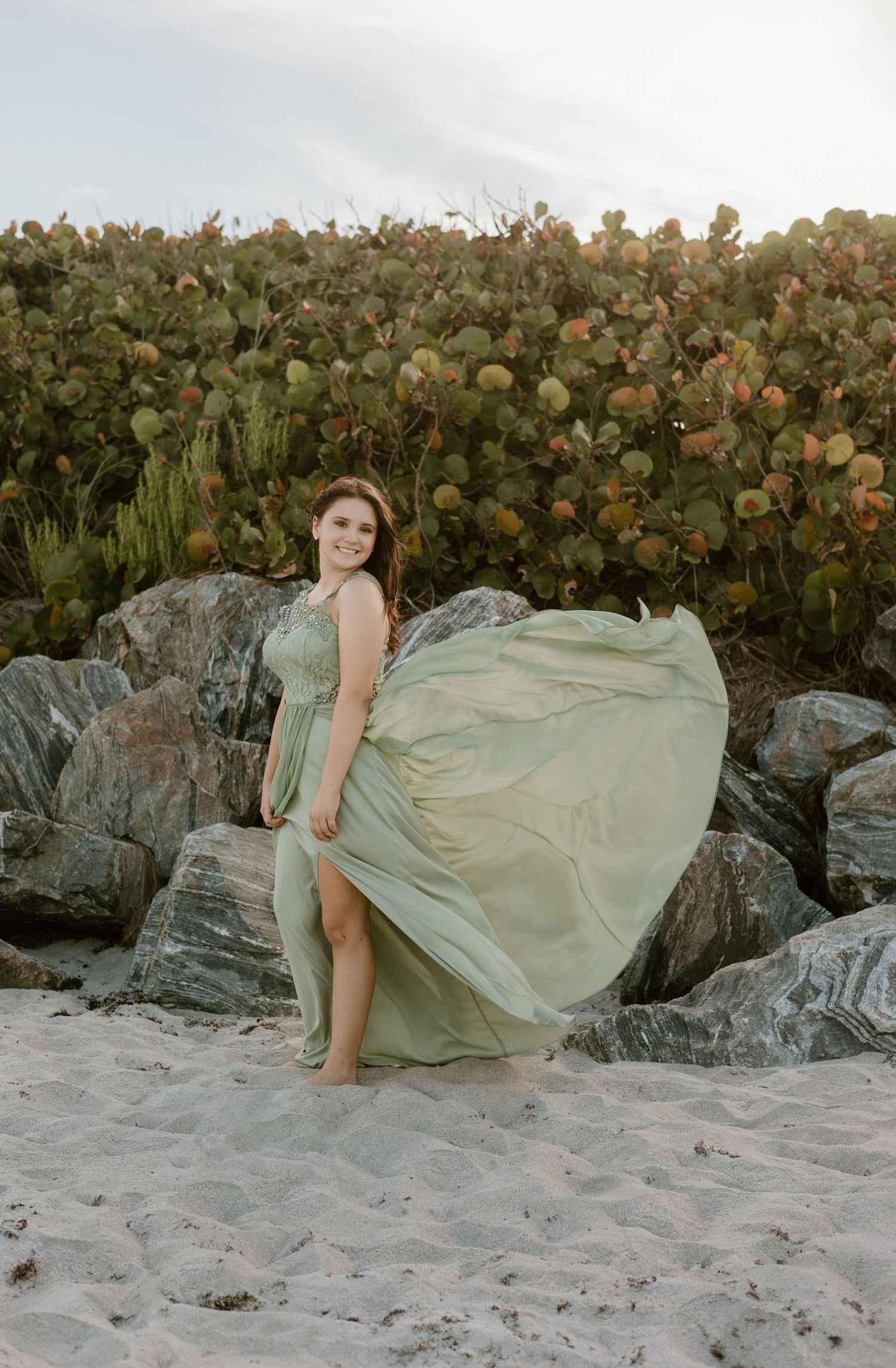 Flowing sage green dress billows in the wind on rocky beach at sunset.