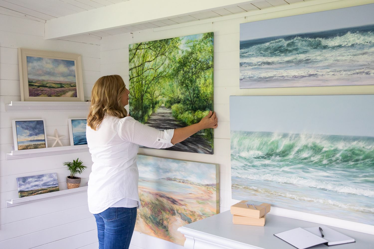 An artist arranges coastal landscape paintings on a white gallery wall during business branding photoshoot.