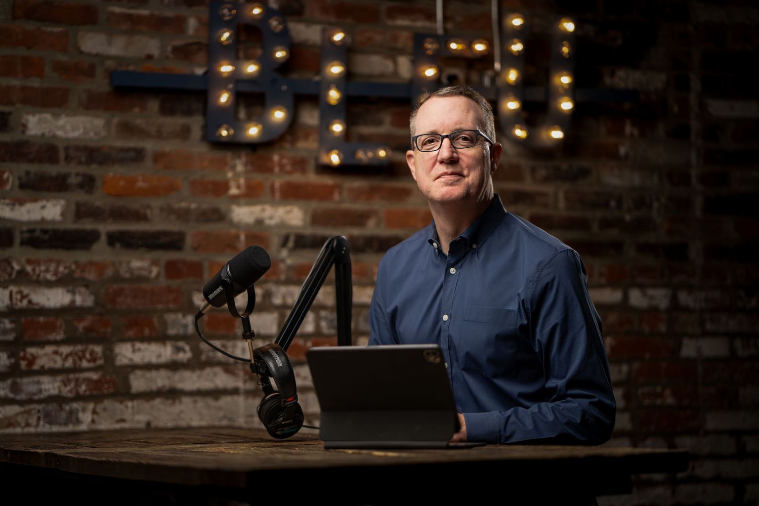 Chris Hill in blue button-down sits at desk with microphone and laptop against brick wall with string lights featured in Boston Magazine
