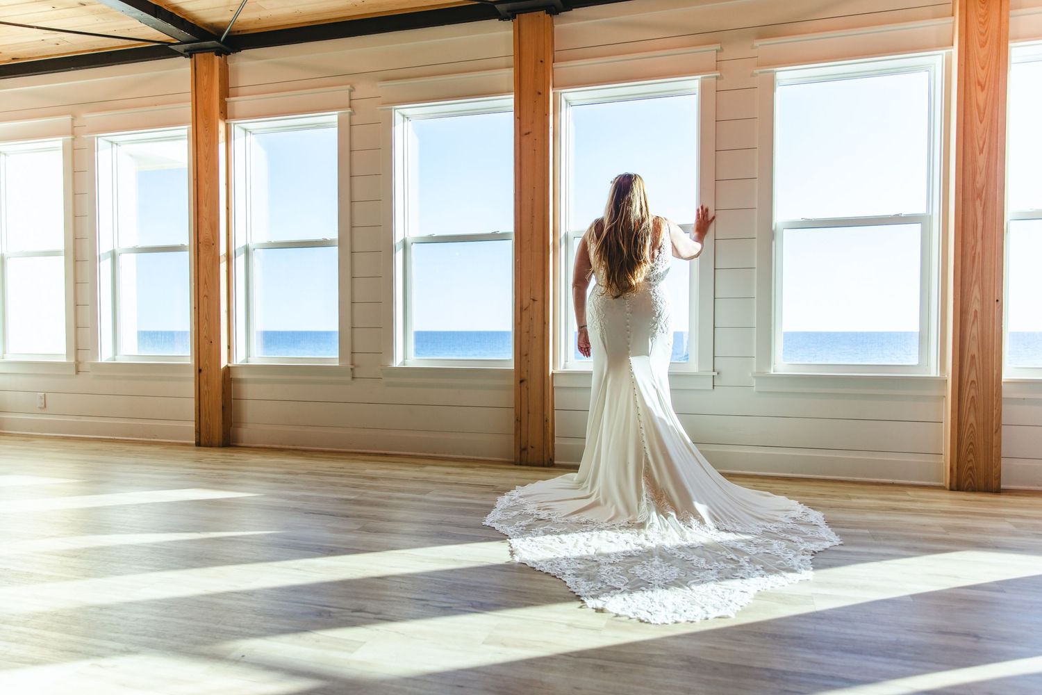 Elegant wedding dress with long train photographed in sunlit room with ocean view windows.