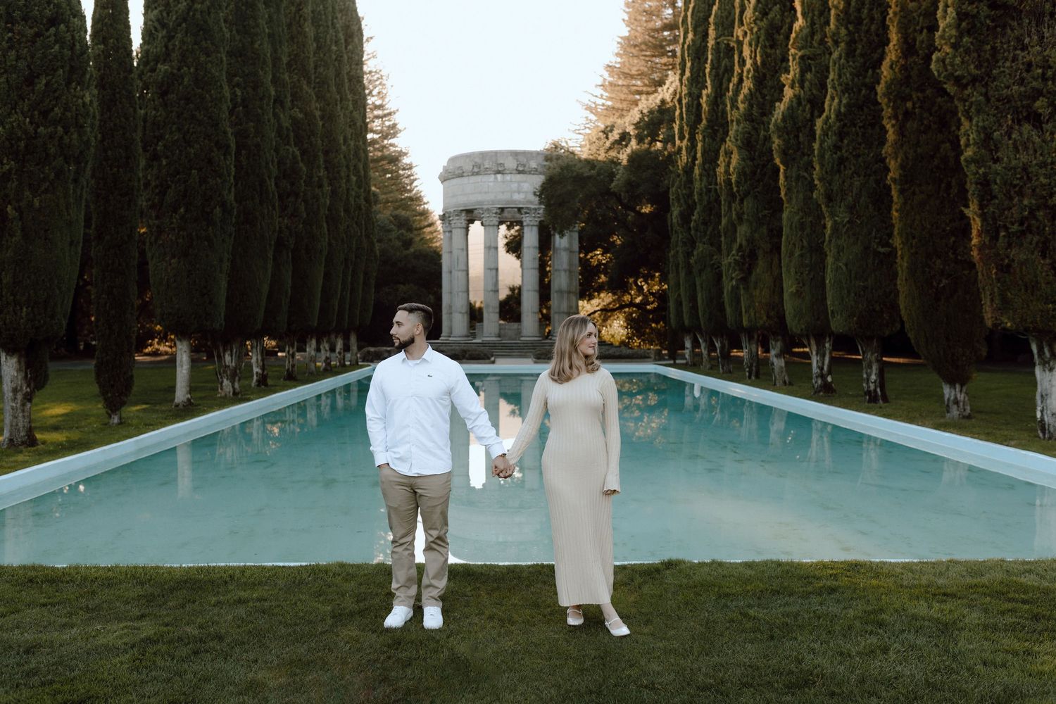 Couple in white outfits standing by reflecting pool surrounded by tall cypress trees.