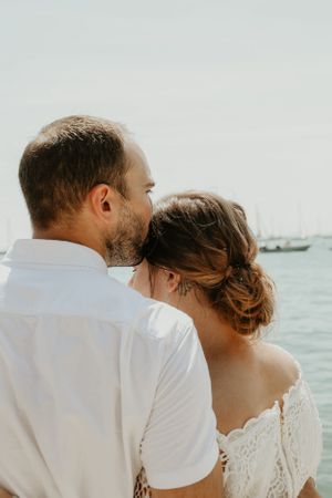 A couple in white clothing embraces by the ocean on a bright day.