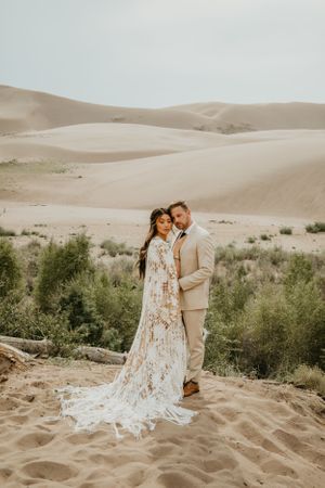 A romantic desert photoshoot featuring a lace dress and beige suit against rolling sand dunes and desert vegetation.