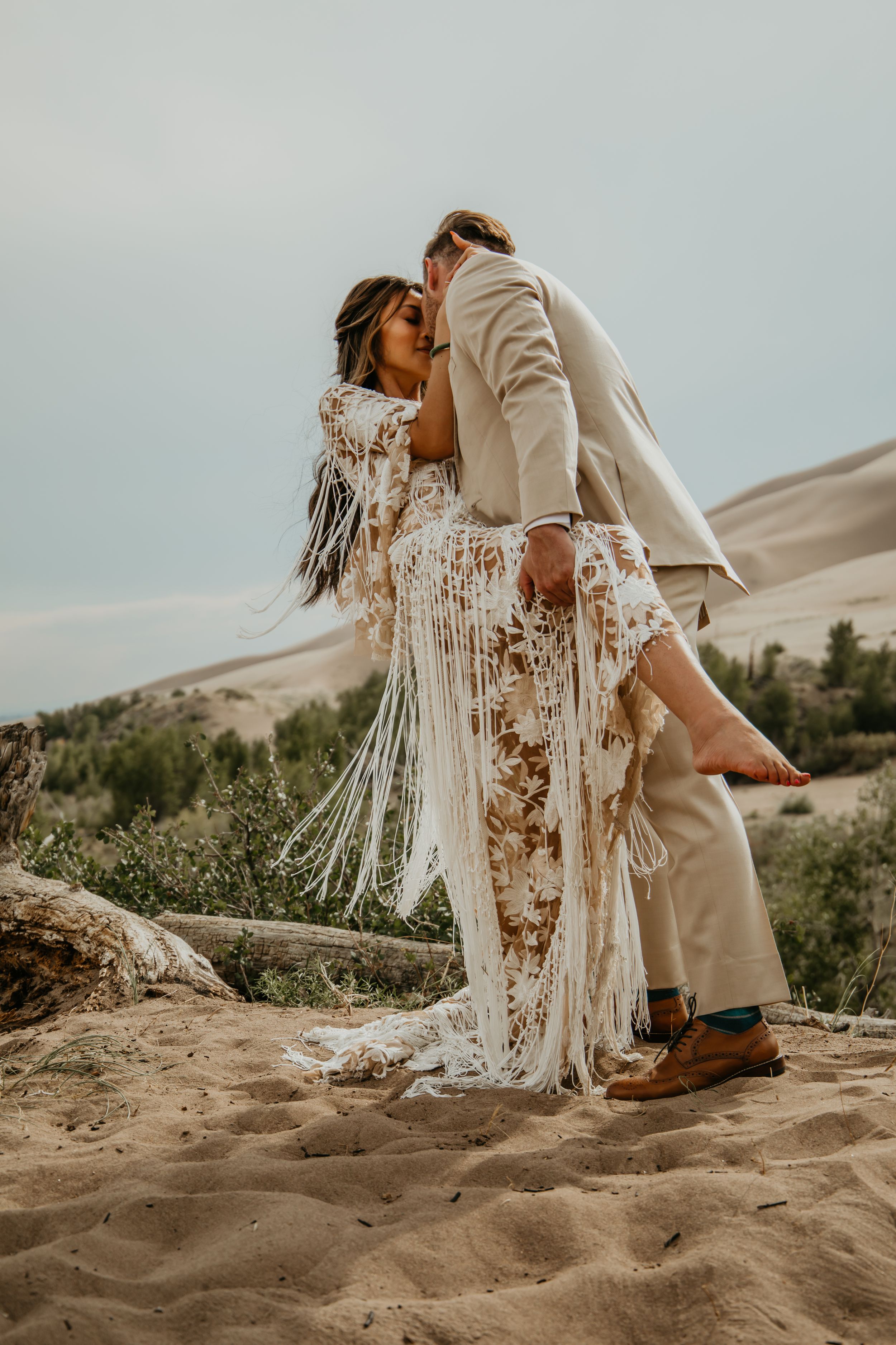 Couple in wedding attire sitting on sandy terrain with desert hills and vegetation in the background.