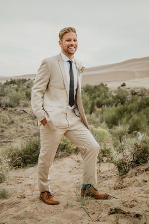 Person in beige suit and brown shoes posing on desert sand with brush in background.