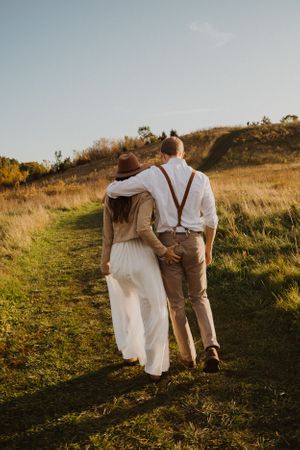 A couple walks hand in hand up a grassy hillside path during golden hour.