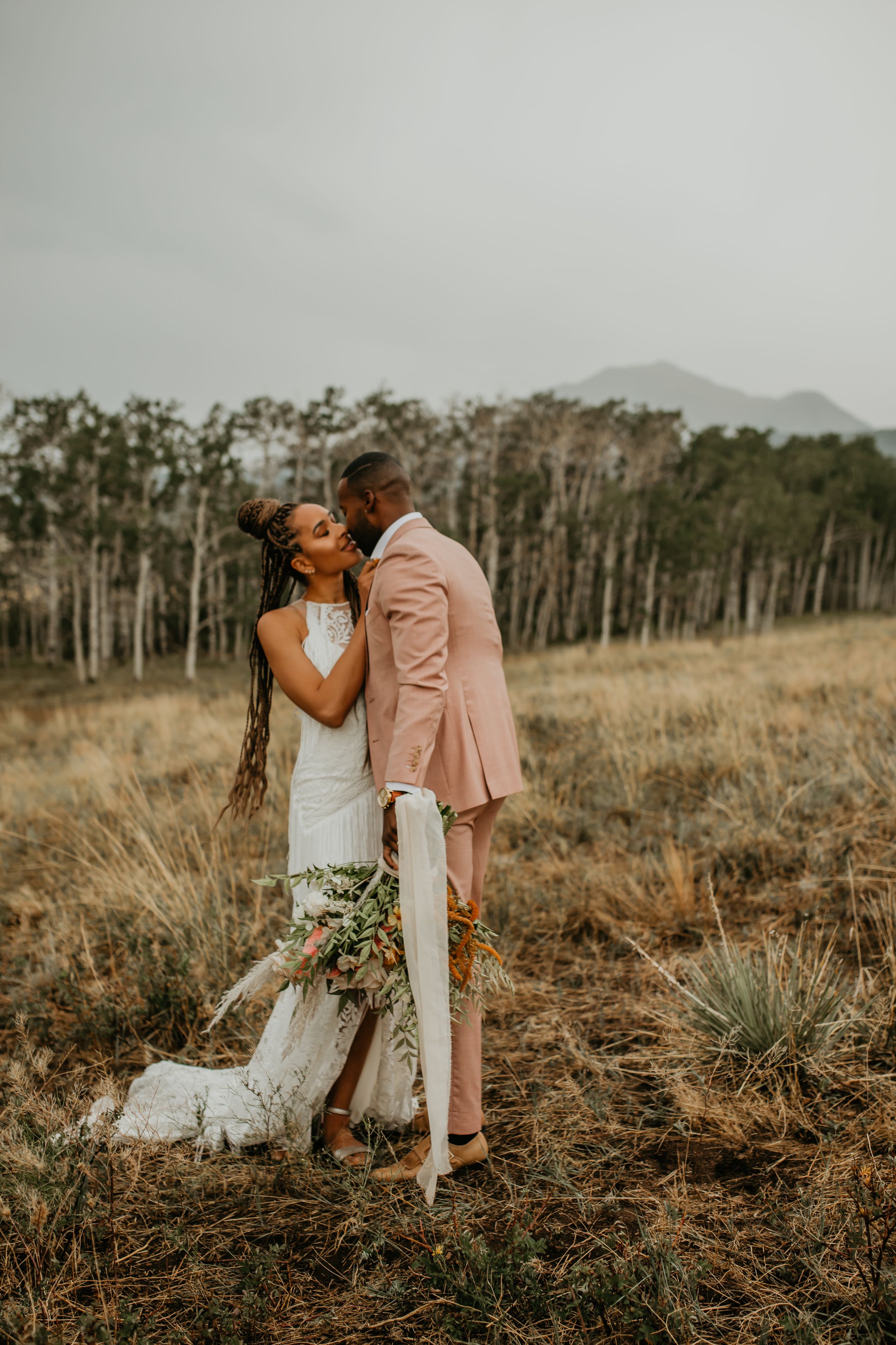 A newlywed couple embracing in a golden field with mountains in the background.