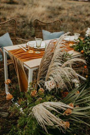 Boho wedding table setting with pampas grass, orange flowers, and wicker chairs in an outdoor setting.