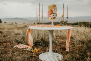 Modern wedding cake display on white pedestal table with tall candles and mountain vista background.