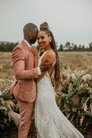 Joyful wedding couple embracing in formal attire against a natural landscape with pampas grass.