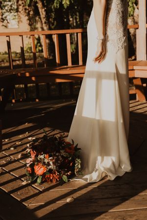 Wedding dress and floral bouquet casting shadows on wooden deck in sunlight.