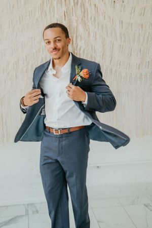 Person in navy suit with orange boutonniere strikes playful poses against white wall.