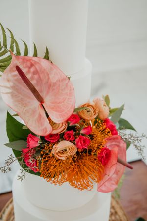 Vibrant tropical floral arrangement with pink anthurium flowers, orange protea, and red roses against a white background.