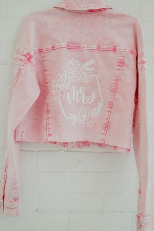 Pink denim jacket with white embroidered details laid flat on white surface.