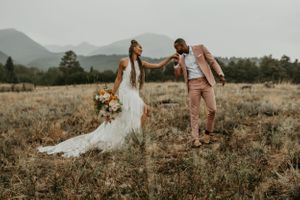 Two people in formal attire stand together in a scenic mountain field at sunset.