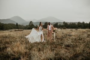 Two figures stand together in a rustic outdoor setting with mountain peaks in the distance.