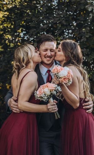 Two bridesmaids in burgundy dresses kiss the groom while holding peach-colored bouquets.