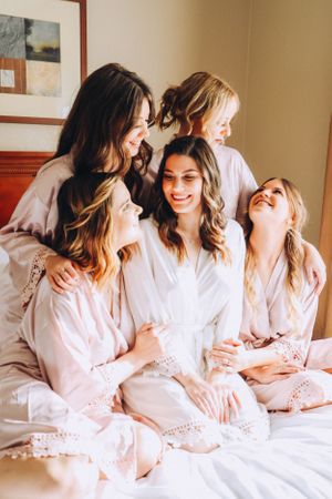 Bridesmaids in pink robes sharing joyful moments on bed during wedding preparations.