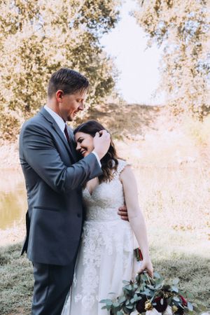 Newlyweds embrace in a golden field during sunset with trees in background.