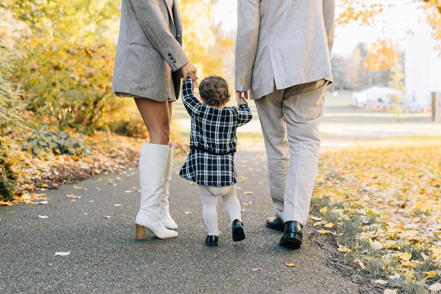 Young family walking toddler between parents on fall foliage lined path.