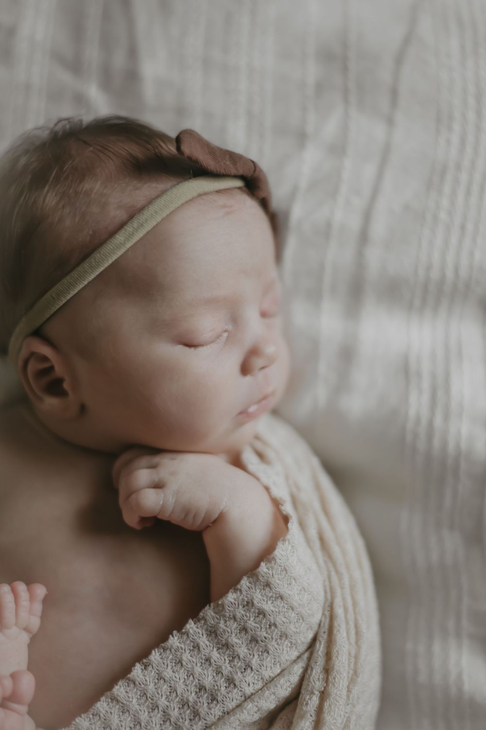 Sleeping newborn baby wrapped in cream knit blanket wearing delicate headband in soft natural lighting.