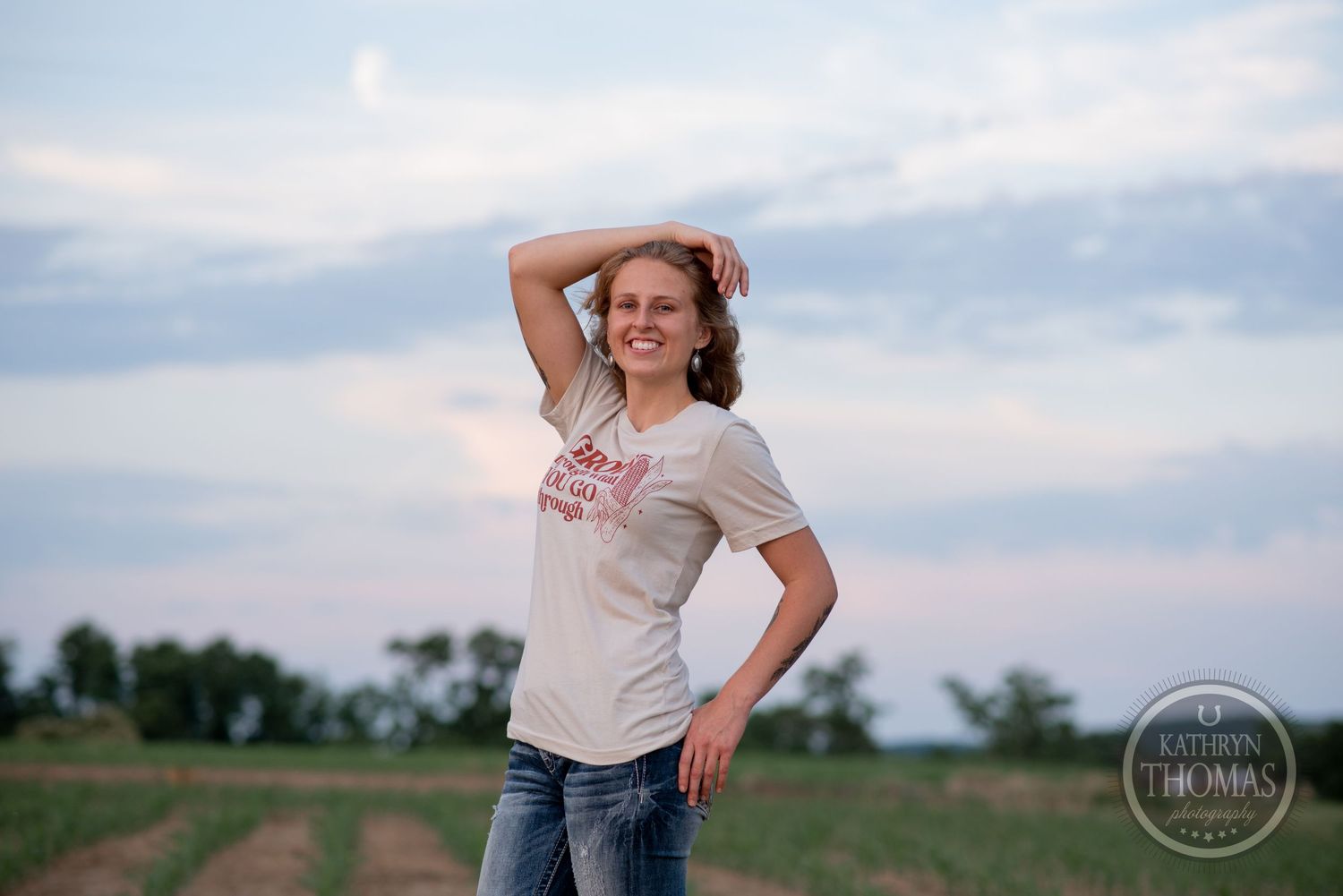 Figure poses in farm field during sunset with cloudy sky backdrop.